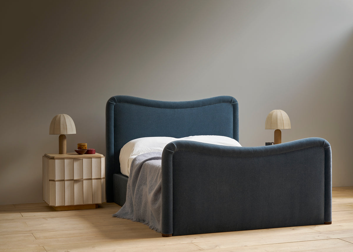 A modern upholstered bed with a curved headboard and footboard in dark blue fabric, styled with white bedding and a gray throw blanket. It is set in a minimalist bedroom with light wood flooring, a matching nightstand with a lamp, and a neutral gray wall.
